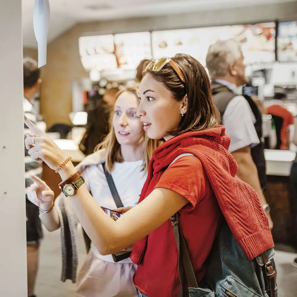 Two friends ordering at a self-service kiosk in a busy quick-service restaurant